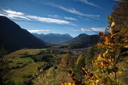 Weitläufiges Tal mit Bergen, herbstlichem Laub und Blick auf eine Ortschaft im Sonnenlicht