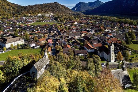 Blick über die Ortschaft Oberau mit einer Kapelle und zwei Kirchen, Herbstbäumen und umliegenden Bergen aus erhöhter Perspektive