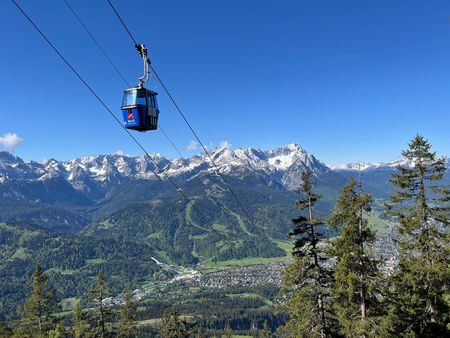 Eine Berggondel über einem Waldgebiet im Hintergrund ein Bergmassiv mit Schnee auf den Gipfeln