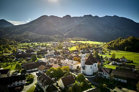 Blick auf ein Dorf mit einer Pfarrkirche. In der Ferne sind grüne Wiesen und bewaldete Berge.