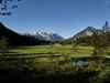 Landschaftsansicht mit grüner Wiese im Vordergrund, Heustadel und Tannen im Mittelgrund und schneebedeckten Bergen im Hintergrund unter klarem, blauem Himmel.
