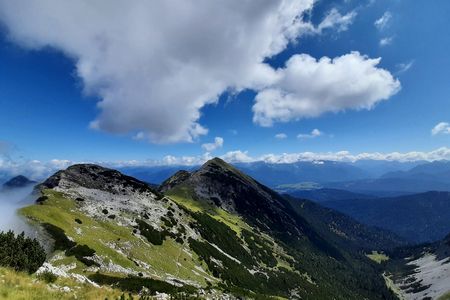 Eine alpine Landschaft mit einem Berggipfel im Vordergrund, grüner Vegetation und bewölktem Himmel. Im Hintergrund sind weitere Bergketten und ein blauer Himmel zu sehen.